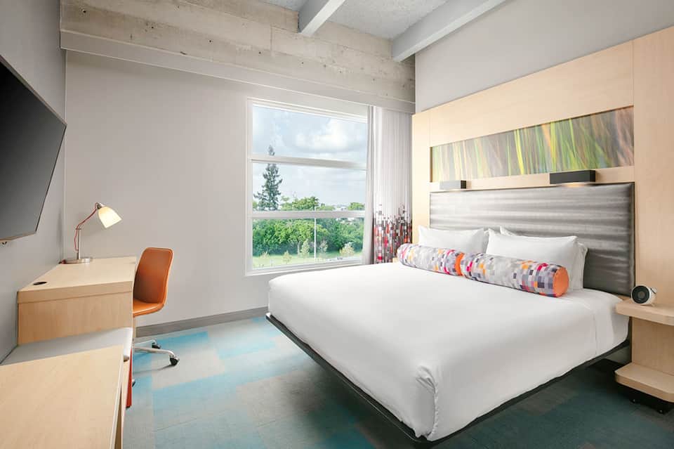 Modern hotel bedroom with queen bed, work desk, floor-to-ceiling windows, and contemporary furnishings