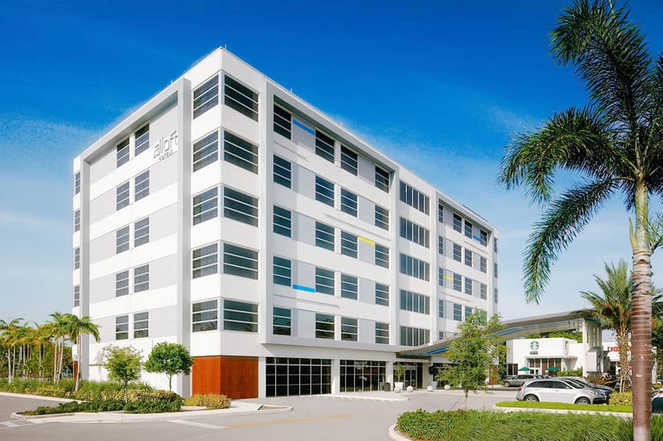 Modern white multi-story hotel building with dark windows and orange accent wall, surrounded by palm trees