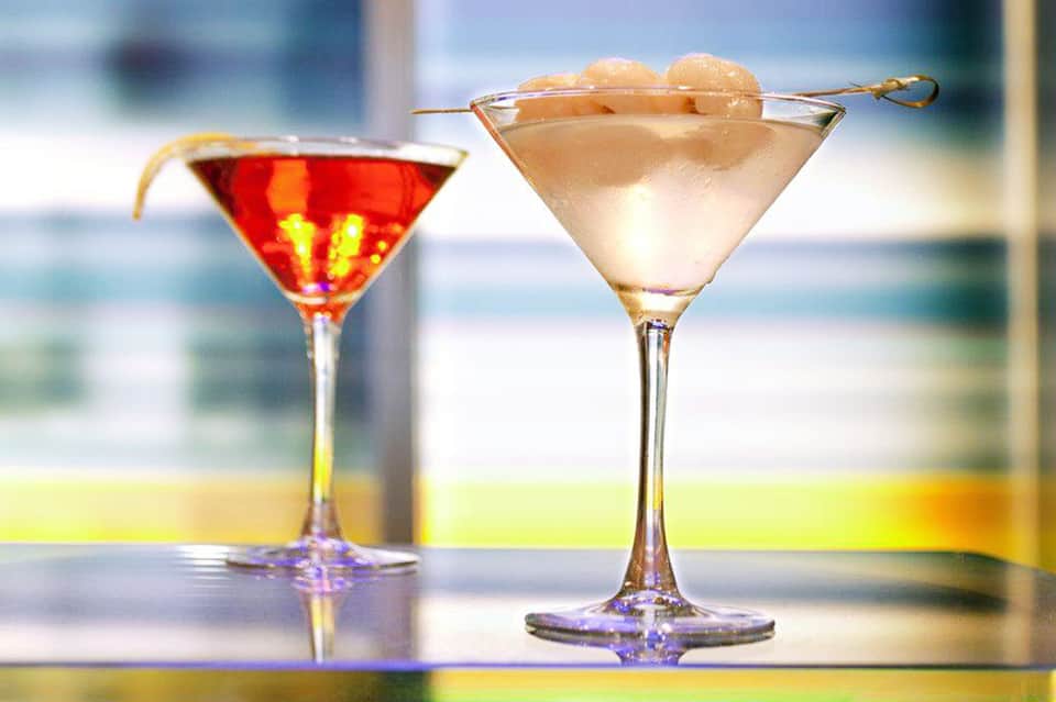 Two colorful cocktails with ice and garnish against blurred ocean and beach backdrop