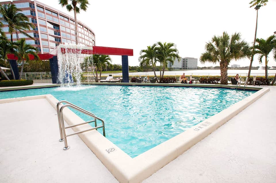 Resort pool with turquoise water, waterfall feature, red umbrellas, and palm trees overlooking waterfront