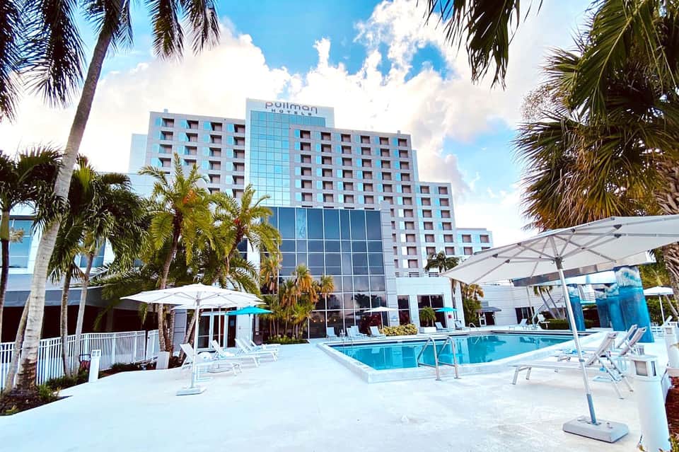 Pullman hotel exterior with pool, white umbrellas, and palm trees on a sunny day