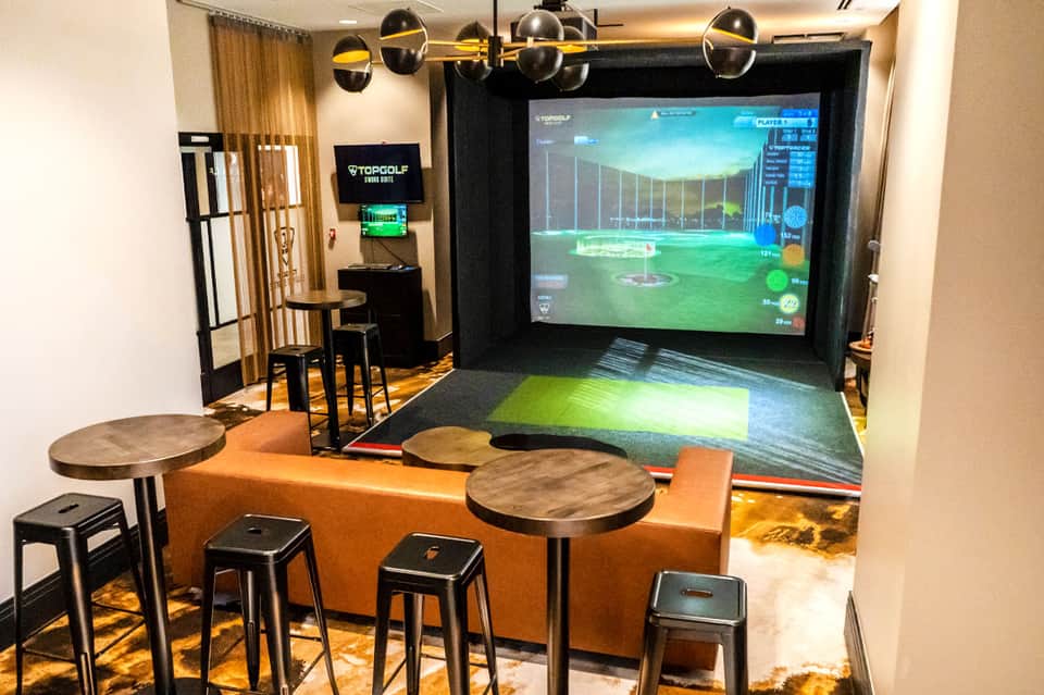 Indoor golf simulator bay with large screen, hitting mat, seating area, and game display