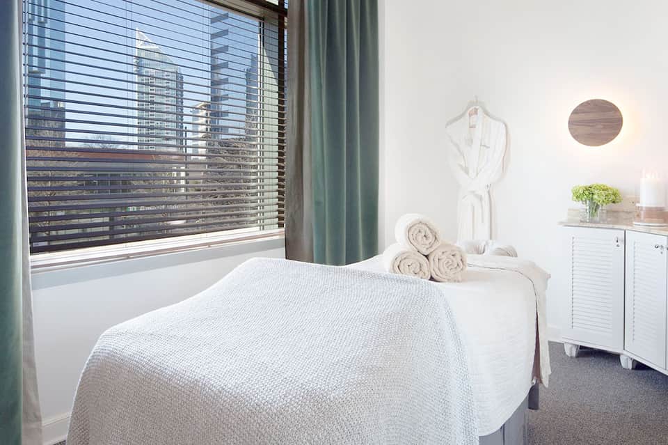 Spa treatment room with massage table, white linens, towels, and window with ocean view