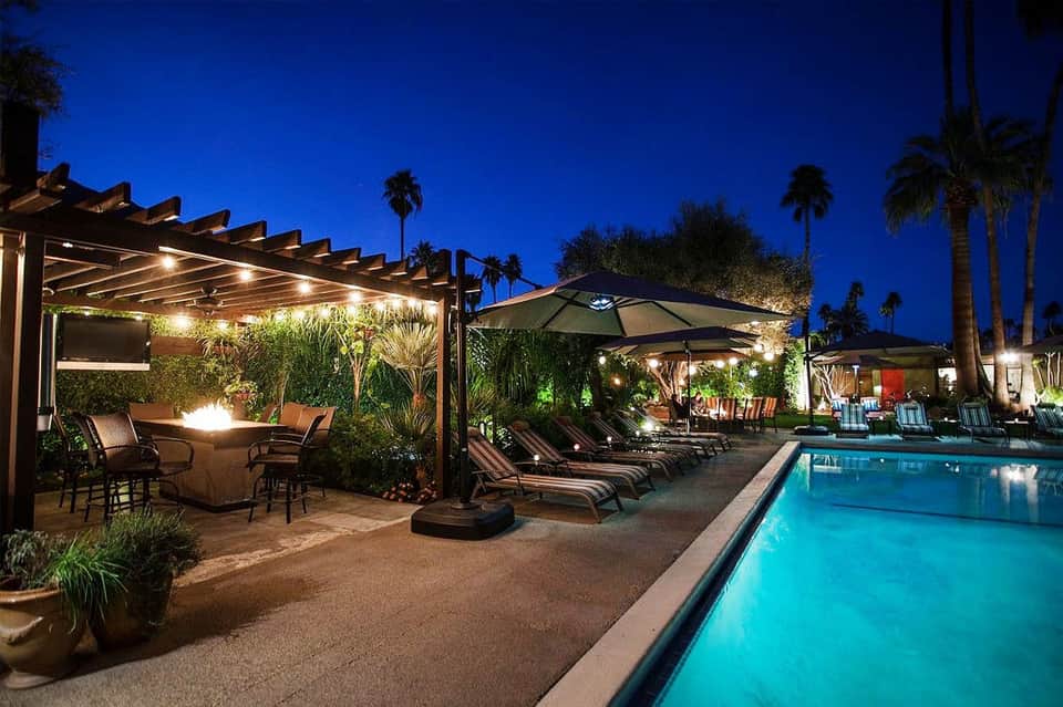 Resort pool at dusk with illuminated pergola, lounge chairs, palm trees, and fire pit