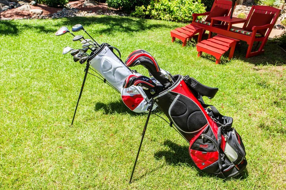 Golf bag with red and black clubs on grass near red Adirondack chairs
