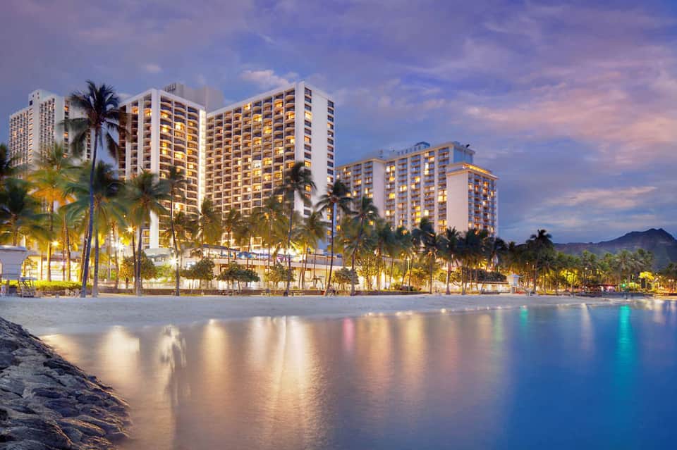 Beachfront resort buildings illuminated at dusk with palm trees and calm ocean waters