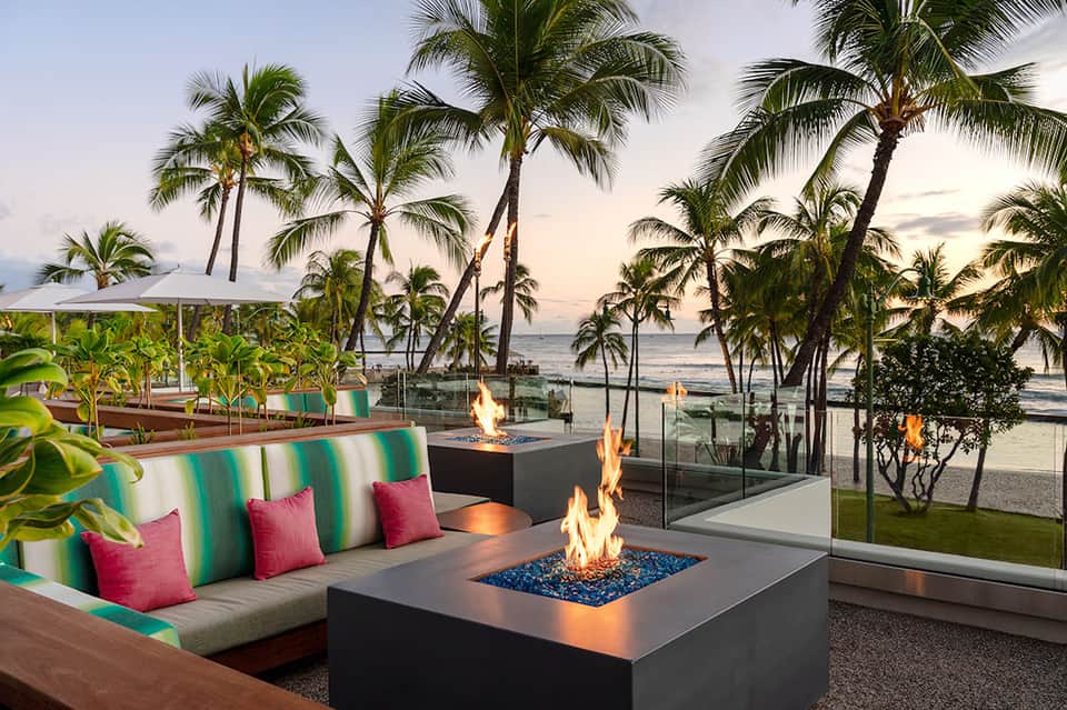 Beachfront lounge area with fire pits, colorful striped seating, palm trees, and ocean sunset view