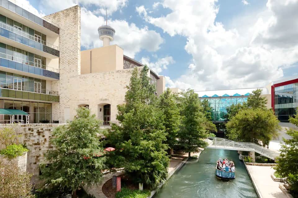 Outdoor urban waterway with boat passing under bridge, surrounded by modern buildings, green trees, and stone architecture.