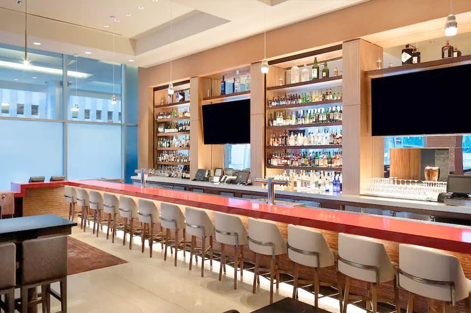 Modern hotel bar with red counter, wooden shelving displaying spirits, and large windows
