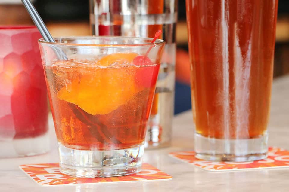 Red cocktail with ice and citrus garnish on patterned coaster with additional drinks