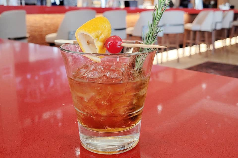 Tropical cocktail with amber liquid, cherry, lemon twist, and rosemary sprig on red bar counter