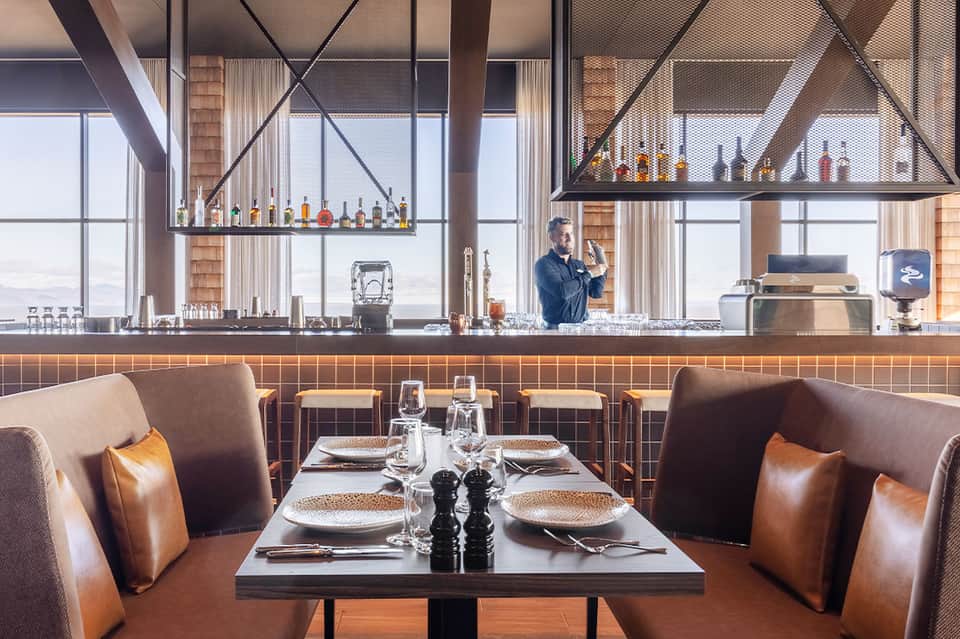 Modern restaurant bar with leather seating, hanging bottle display, and ocean views