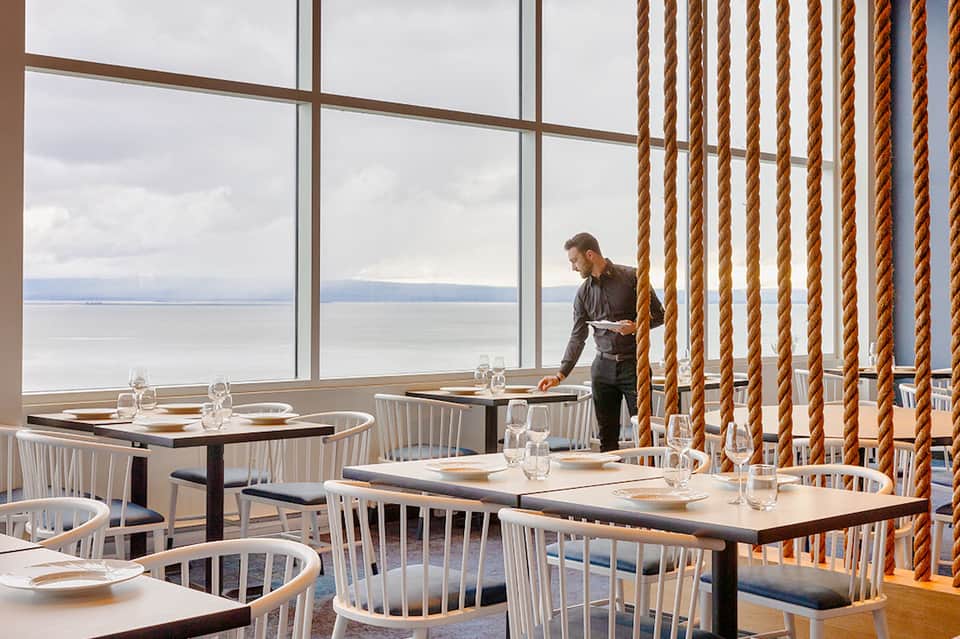 Modern restaurant dining area with floor-to-ceiling windows overlooking calm ocean waters and rope curtains