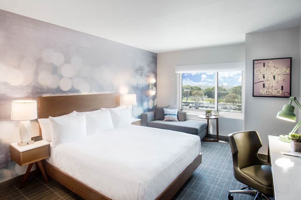 Contemporary hotel bedroom with queen bed, sitting area, and panoramic windows overlooking grounds