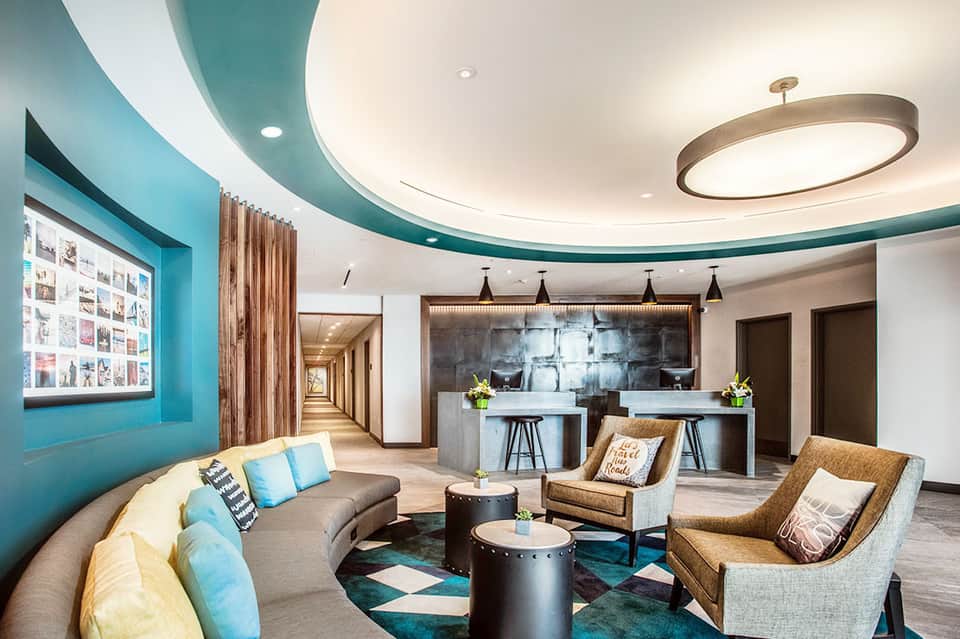 Modern hotel lobby lounge with turquoise accent walls, curved sofa, bar area, and contemporary lighting