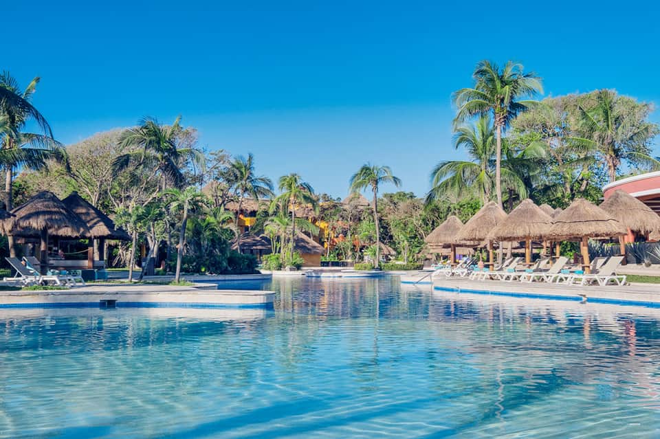 Resort pool with thatched-roof cabanas, palm trees, and ocean views