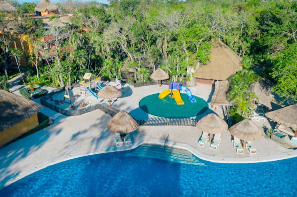 Aerial view of resort with lagoon-style pool, play area, and thatched-roof structures surrounded by trees