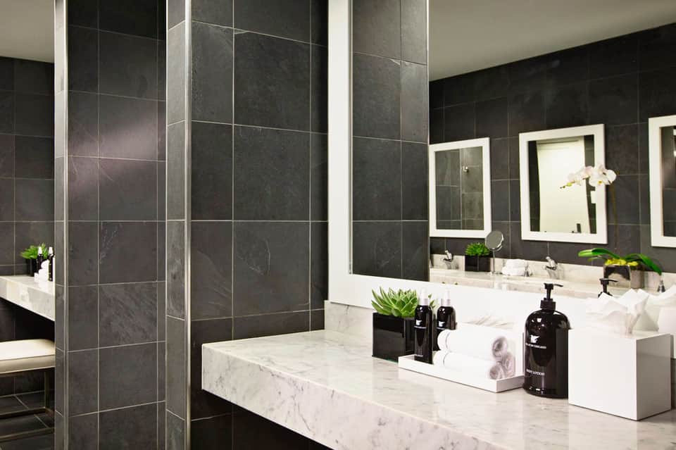 Luxurious bathroom with dark tile walls, marble countertop, white vessel sinks, and illuminated mirrors
