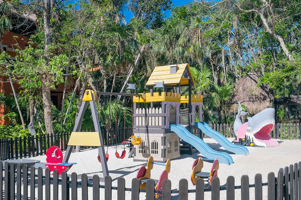 Children's playground with yellow and gray castle structure, blue slides, and swings in shaded area