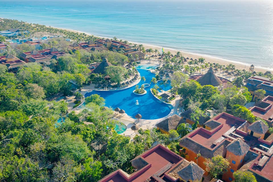 Aerial view of beachfront resort with blue lagoon pool, thatched-roof buildings, and palm trees