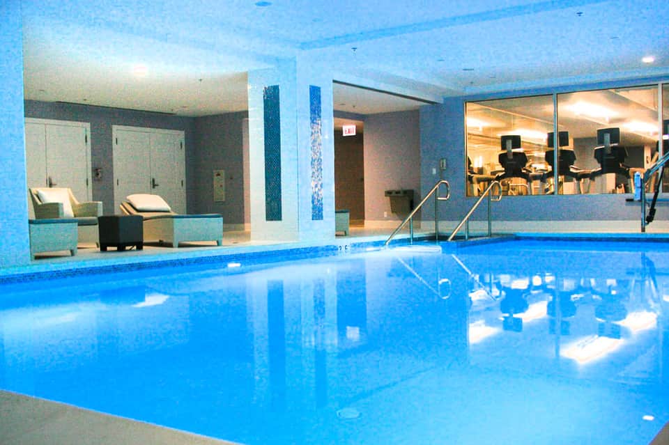 Indoor swimming pool with blue tiled walls, lounge chairs, and a fitness center visible through glass windows