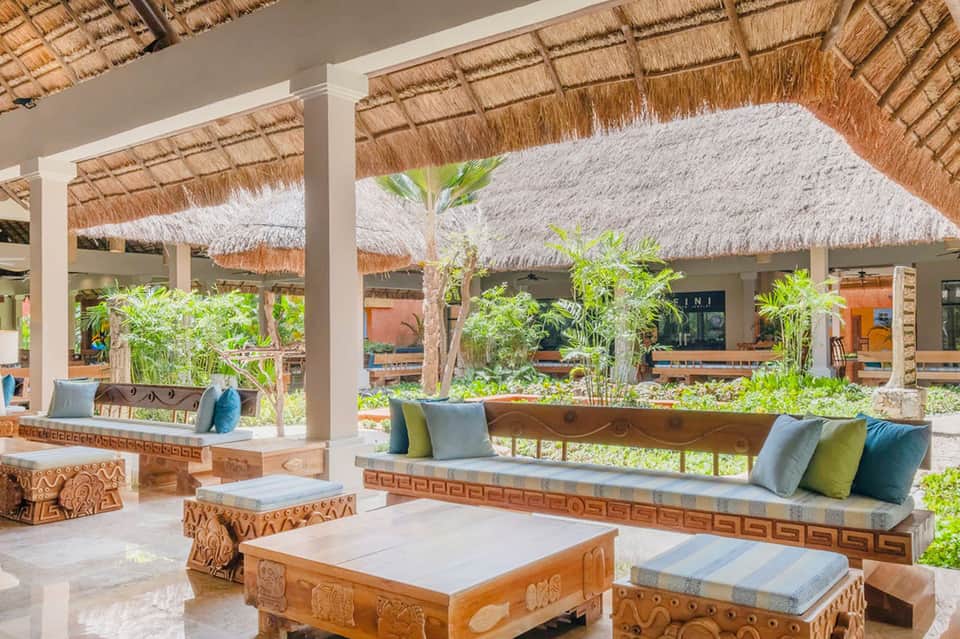 Beachfront resort lounge area with thatched roof, wooden furniture, and tropical plants