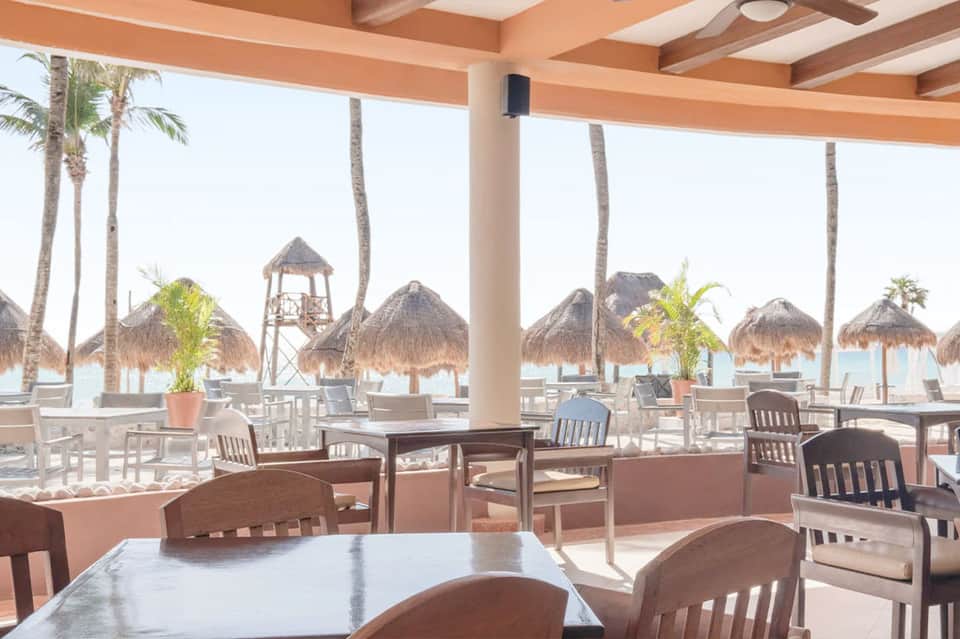 Beachfront restaurant dining area with ocean view, palm trees, thatched umbrellas, and casual seating