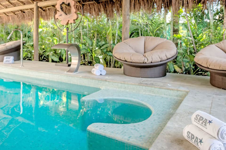 Resort pool deck with lounge chairs, palm trees, and thatched-roof structures in tropical setting