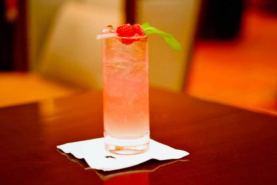 Coral-colored cocktail with ice, raspberries, and mint garnish on a white coaster