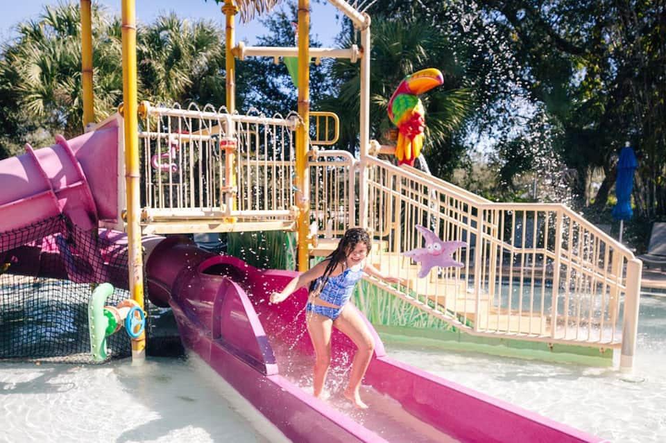 Children's water playground with colorful pink slide, white railings, and tropical palm trees