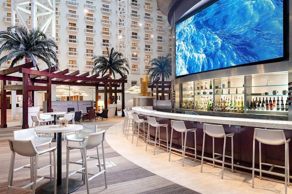 Modern resort bar lounge with high ceilings, purple pergola, large water display screen, and full liquor selection