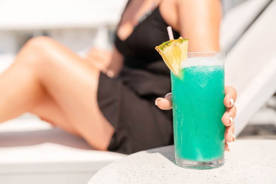Turquoise tropical cocktail with pineapple garnish held by person in black swimsuit