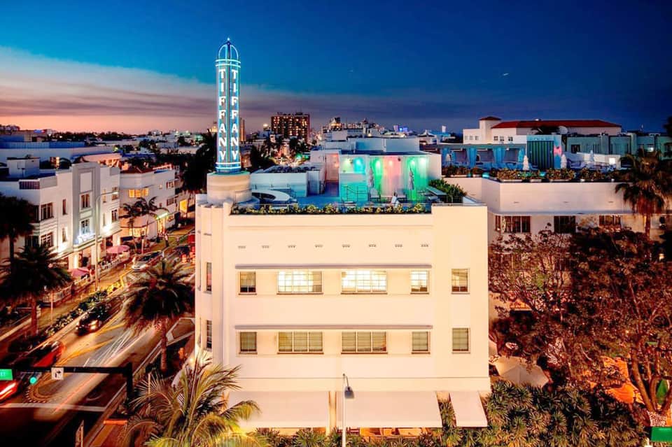 Rooftop bar and lounge area at dusk with colorful lighting, palm trees, and city skyline views