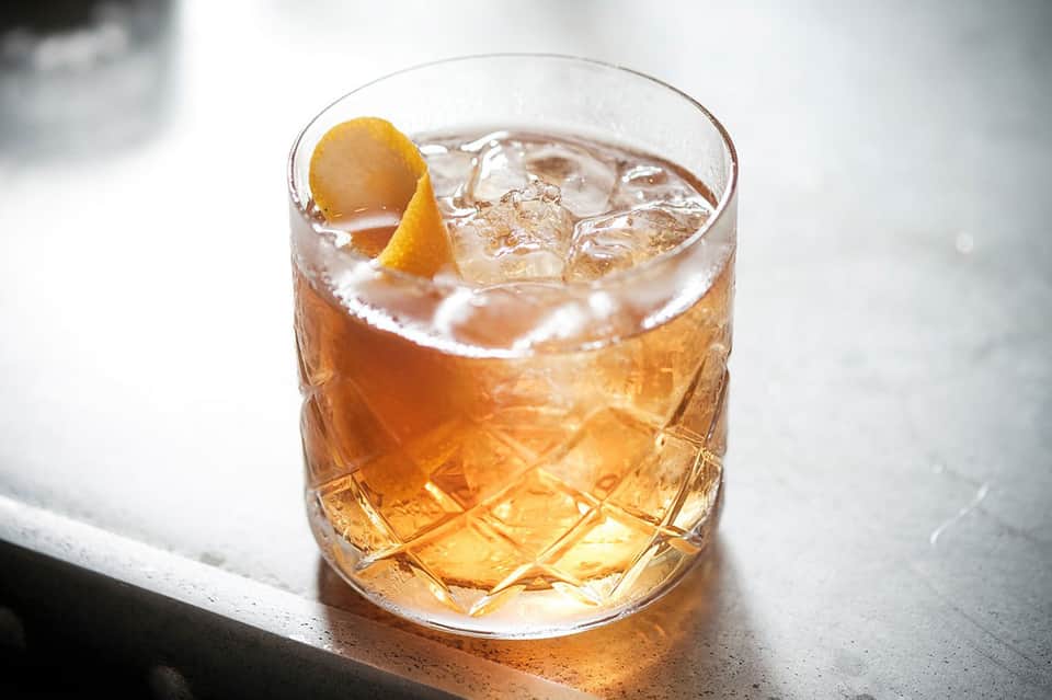 Amber cocktail with ice and orange peel garnish on white surface