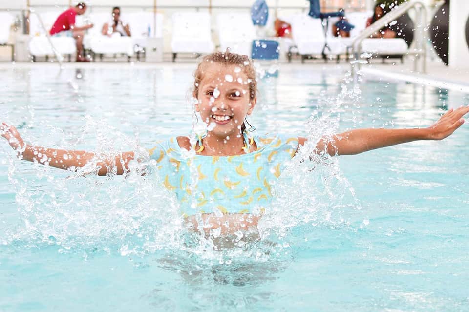 Young girl splashing joyfully in shallow pool water wearing swim float