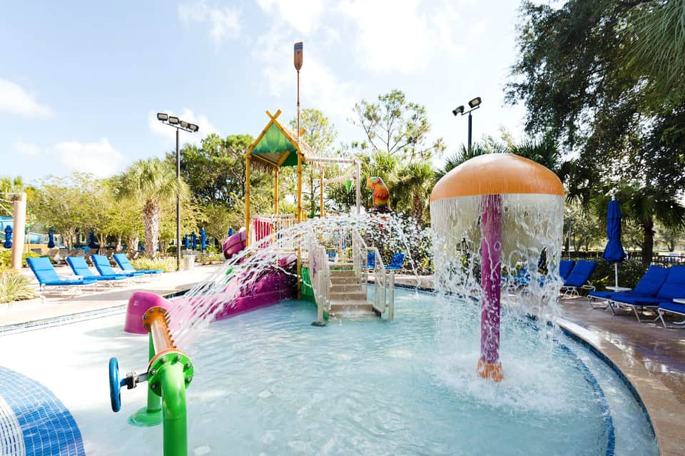 Colorful outdoor water park with splash pad, slides, and play structures surrounded by palm trees and lounge chairs