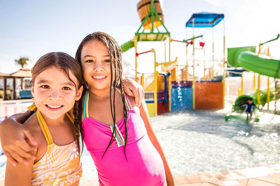 Two young girls in swimsuits smiling at colorful water park with slides and splash pad