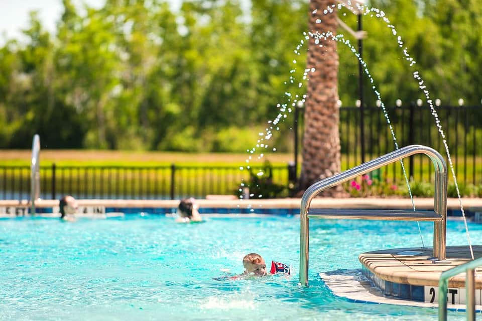 Child swimming in resort pool near metal ladder with palm trees and manicured lawn