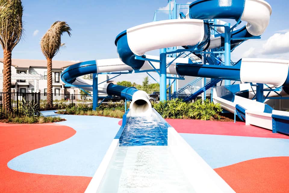 Water park with blue and white slide structures on red and blue padded surface near white residential buildings