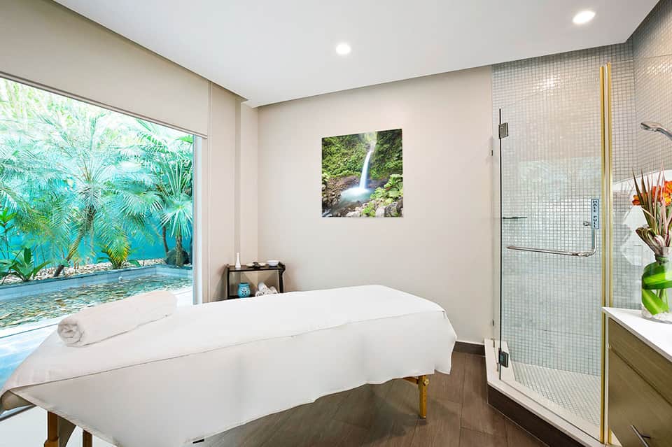 Spa treatment room with soaking tub overlooking tropical garden through large window and modern shower