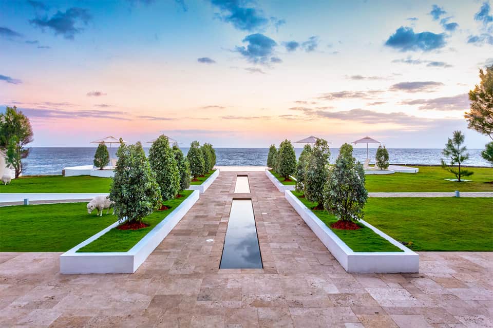 Elegant seaside promenade with reflecting pool, manicured lawns, palm trees, and ocean sunset backdrop