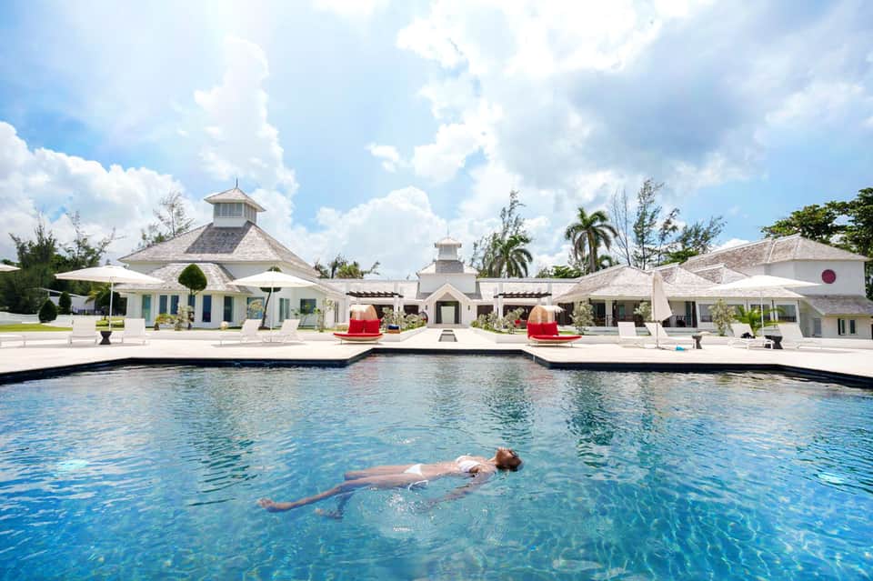Resort pool with person swimming, surrounded by white cabanas and tropical palm trees under blue sky