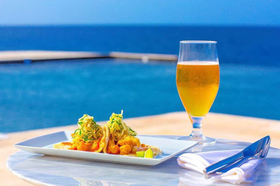 Gourmet plated dish and beer glass on table overlooking blue ocean and infinity pool