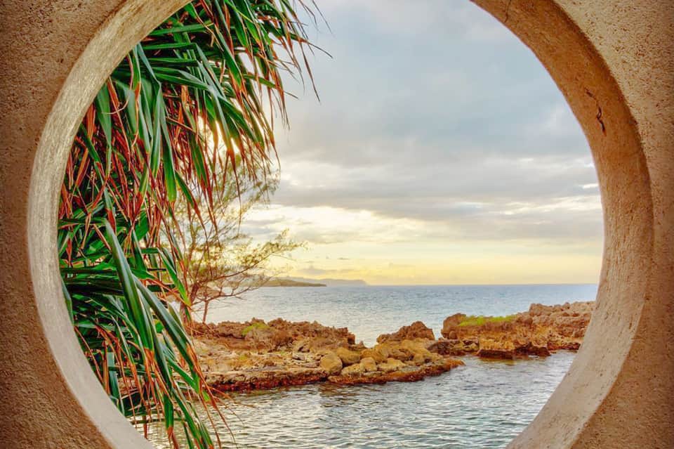 Circular concrete archway framing ocean view with tropical plants and rocky coastline at sunset