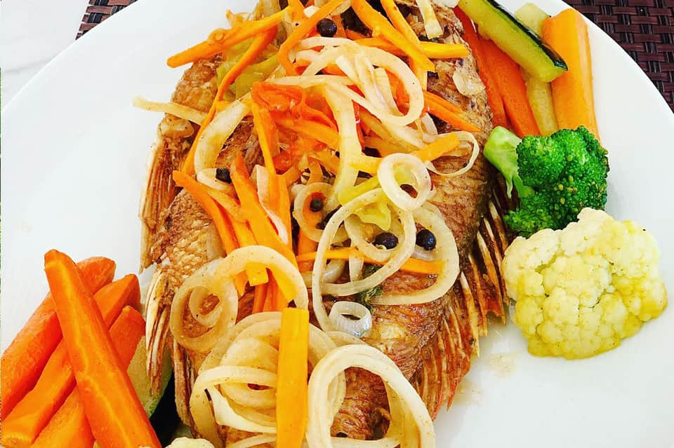 Grilled fish fillet topped with sautéed onions and peppers, served with colorful vegetables on white plate