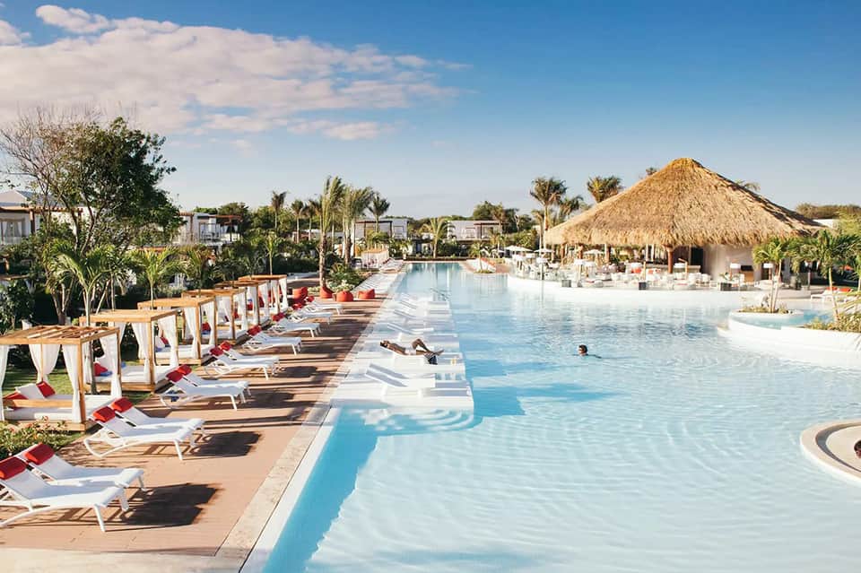 Resort pool with cabanas, lounge chairs, and thatched roof structure surrounded by palm trees