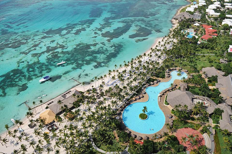 Aerial view of tropical beach resort with curved pool, white buildings, palm trees, and turquoise ocean