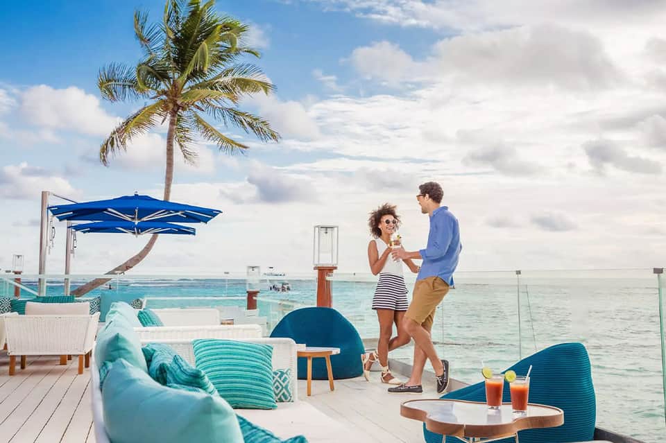 Couple with drinks on beachfront deck with palm tree and blue umbrella