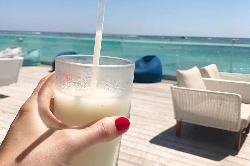 Hand holding cold beverage with straw overlooking turquoise ocean and white lounge chairs