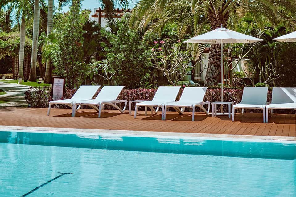 Resort pool deck with white lounge chairs, umbrellas, tropical palms, and lush landscaping